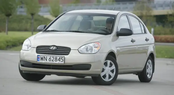 Złoty Hyundai Accent z kierowcą za kierownicą, zaparkowany na zewnątrz.
