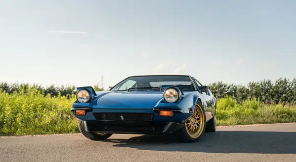 Niebieski De Tomaso Pantera z wysuwanymi światłami i złotymi felgami na tle zieleni i błękitnego nieba.
