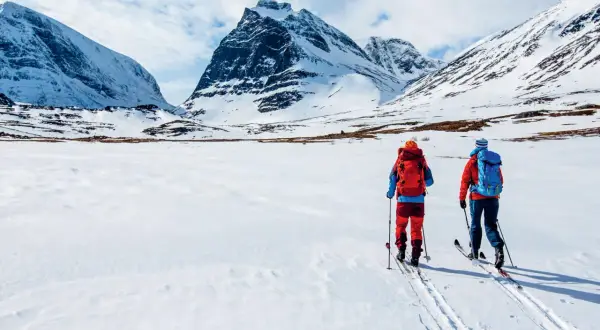 Två skidåkare med ryggsäckar på väg att bestiga berg i snöklädda fjäll under en molnig himmel.