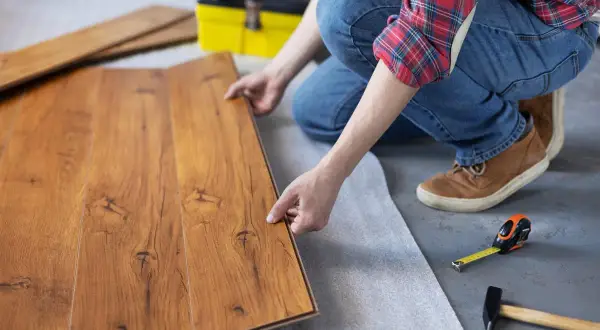 Man instalando suelo de tarima flotante. Se ven las lamas de madera, una cinta métrica y un martillo.