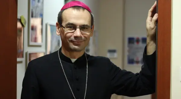 Uśmiechnięty, najmłodszy biskup w Polsce, w czarnej sutannie i z różowym zucchetto na głowie, opiera się o framugę drzwi.