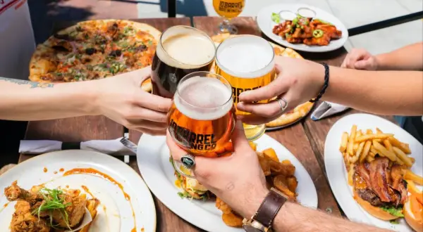 Ludzie wznoszą toasty piwem, celebrując spożycie piwa na świecie przy stole pełnym pizzy, burgerów i kurczaka z goframi.