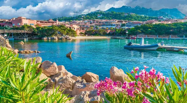 Vue d'une baie turquoise en Sardaigne, avec des maisons roses, un pont en bois et des fleurs roses. Idéal pour savoir que faire en Sardaigne.