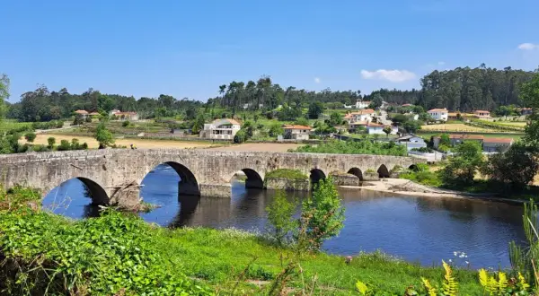 El ponte de Outeiro, un puente de piedra con varios arcos, cruza un río azul. En la orilla opuesta, casas con tejados rojos se asientan en una colina verde.