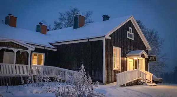 Mörkt trähus med snö på taket och runt om. Varmt ljus lyser från fönster och entré. Skylt med texten "Stilleben" syns.