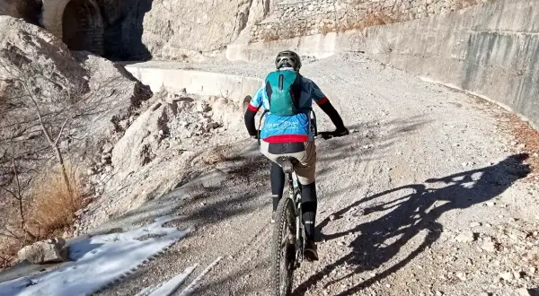 Ciclista in mountain bike su sentiero sterrato, con neve residua e un tunnel scavato nella roccia. Un'avventura tra le **Tignale cascate**.
