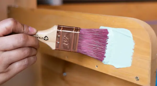 Holzbank streichen ohne schleifen: Ein Pinsel mit hellblauer Farbe wird über das Holz geführt.