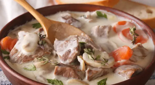 Un plat réconfortant de véritable recette blanquette de veau, avec des morceaux de viande tendre, des champignons et des carottes dans une sauce crémeuse, servi dans un bol en bois avec une cuillère.
