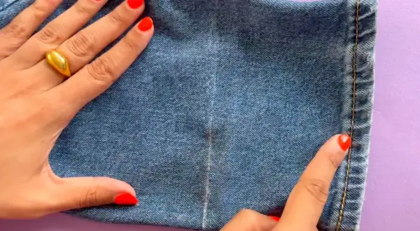 Main d'une femme avec un ongle rouge vif et une bague dorée, ajustant l'ourlet d'un jean.