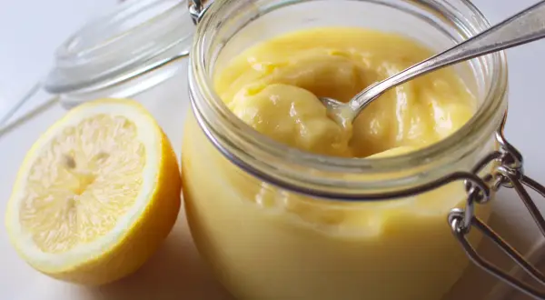 En burk med krämig lemon curd och en halv citron bredvid. En sked vilar i burken, redo att skopa upp den syrliga godsaken.