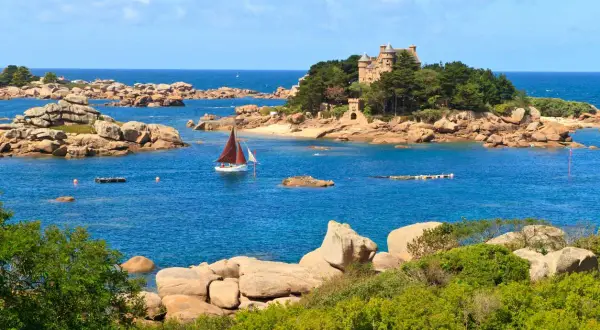 Un voilier navigue près d'un château sur une île rocheuse en Bretagne. Le paysage est typique de la côte bretonne.