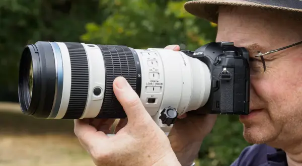 A photographer in a hat uses a large telephoto lens on a Micro Four Thirds camera, perhaps for a detailed micro 4/3 lenses review.