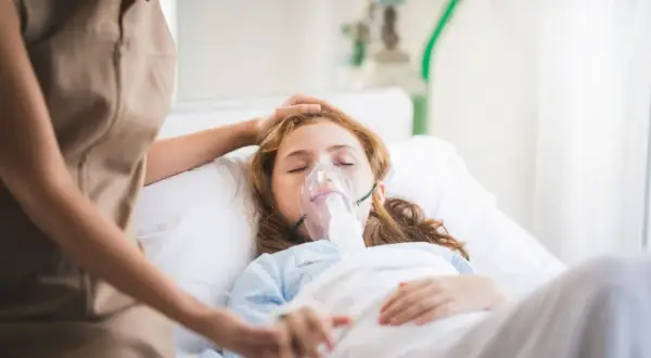 Dziewczynka w szpitalnym łóżku z maską tlenową, walcząca z hipoksją. Opiekunka czule kładzie dłoń na jej głowie.