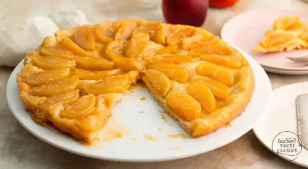 Eine goldbraune Tarte Tatin mit karamellisierten Apfelspalten auf einem weißen Teller. Ein Stück fehlt, was Appetit macht.