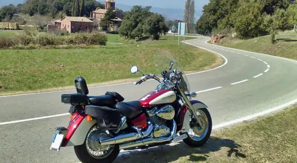 Honda VT 750 C Shadow 2007 ficha técnica. Moto custom roja y crema aparcada en una carretera rural con un paisaje de colinas y edificios antiguos.