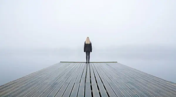 Una persona fredda, con la psicologia che la isola, sta in piedi su un molo di legno in una giornata nebbiosa.