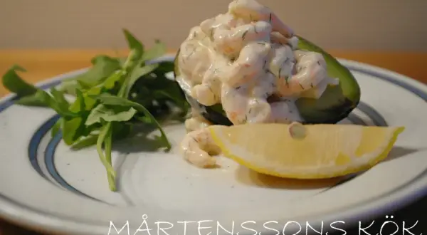 Avokadohalva fylld med räkor i en krämig sås, liknande guacamole crème fraiche, med en citronskiva och gröna blad.