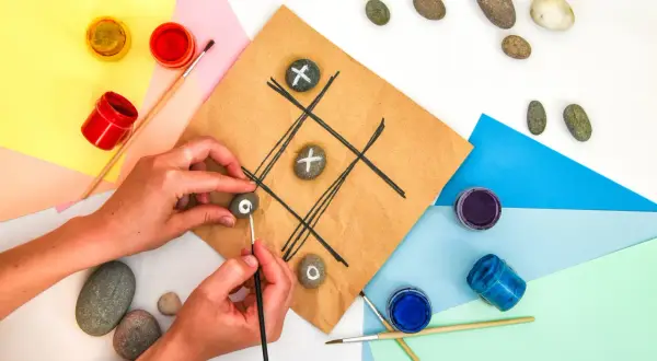 Mani che creano un gioco del tris fai da te dipingendo simboli su sassi. Materiali colorati e pennelli completano la scena.