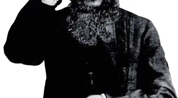 Grigorij Rasputin, z charakterystyczną brodą, wznosi rękę w geście błogosławieństwa lub pozdrowienia.