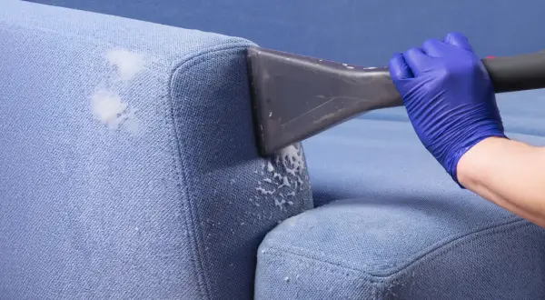 Blaue Polster reinigen mit Rasierschaum. Ein Handschuh hält ein Reinigungsgerät, das Schaum auf den Stoff sprüht.
