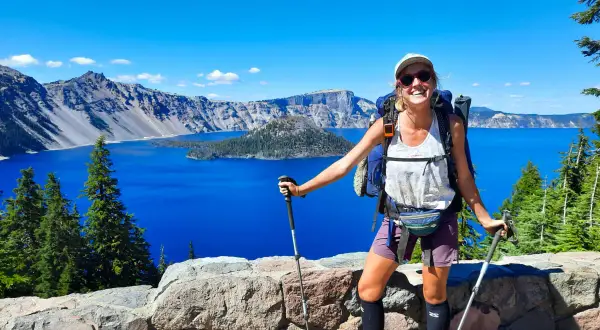 Uśmiechnięta turystka z plecakiem i kijkami, podziwiająca błękitne jezioro. To widok z trasy Pacific Crest Trail, pokazujący, ile się idzie przez takie cuda natury.