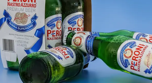 Butelki piwa Peroni Nastro Azzurro leżą na niebieskim tle. Jedna z butelek jest otwarta, a pianka unosi się z szyjki.