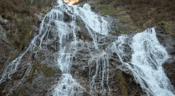 La cascada de Aitzondo desciende por una ladera rocosa, con agua blanca y espumosa que se abre paso entre la vegetación seca y las rocas grises.
