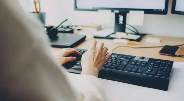 Dłonie piszą na klawiaturze, za nimi monitor. Szybkie załatwienie sprawy, może zwolnienie lekarskie online?