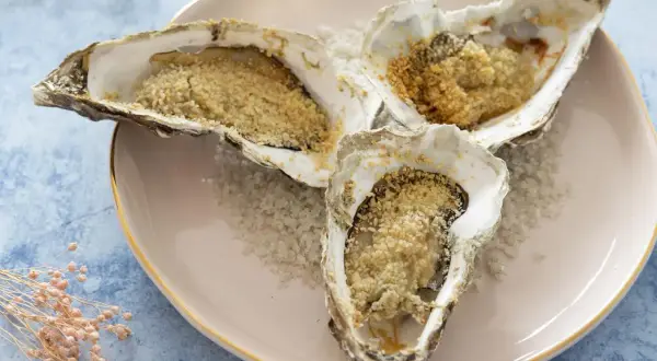 Trois huitres au four, garnies de chapelure dorée, reposent sur un lit de gros sel sur une assiette rose.