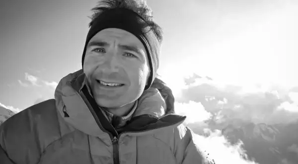 Ueli Steck, lächelnd in warmer Bergkleidung, blickt in die sonnige, wolkenverhangene Bergwelt.