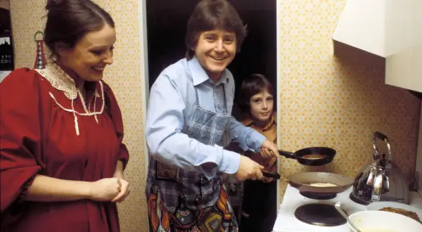 Familie in der Küche, der Vater brät Pfannkuchen nach DDR Rezepten. Ein Kind schaut zu, die Mutter lächelt.