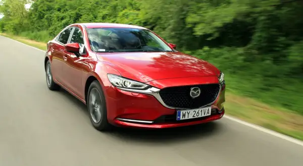 Czerwona Mazda 6 2.0 benzyna pędzi po wiejskiej drodze, otoczona zielenią drzew.