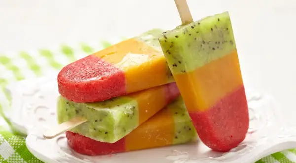 Kolorowe, warstwowe domowe lody na patyku z kiwi, mango i truskawkami, ułożone na białym talerzyku.