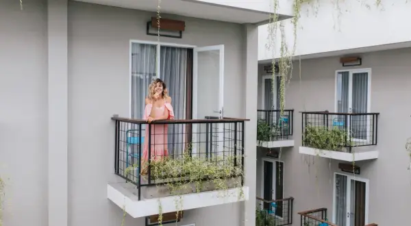 Kobieta w różowej sukience stoi na balkonie, opierając się o balustradę. Wokół rosną zielone rośliny, tworząc naturalną zasłonę na balkon.