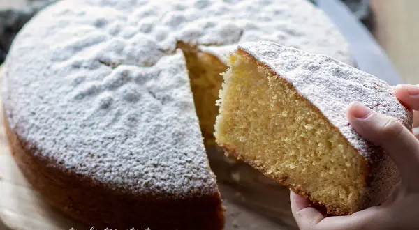 Una fetta di torta margherita soffice, cosparsa di zucchero a velo, viene sollevata da una mano.