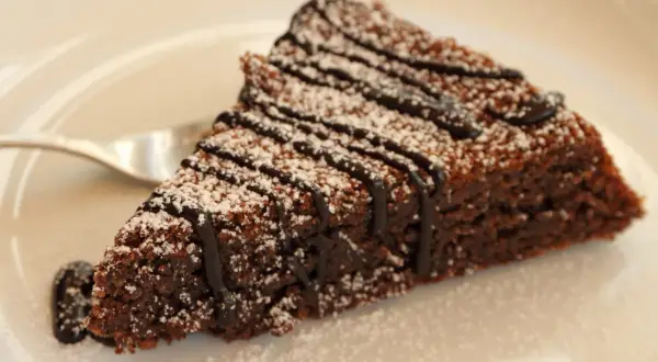 Una fetta di torta caprese al cioccolato, cosparsa di zucchero a velo e con una colatura di cioccolato fondente.