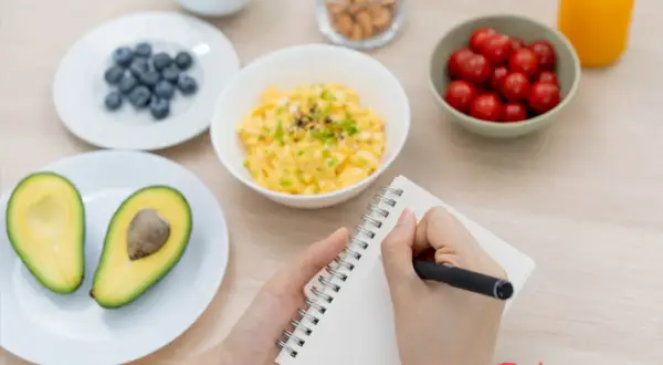 Manos escribiendo en un cuaderno, rodeadas de ingredientes para crear ejemplos de menú: aguacate, arándanos, huevos revueltos, tomates cherry, frutos secos y jugo.