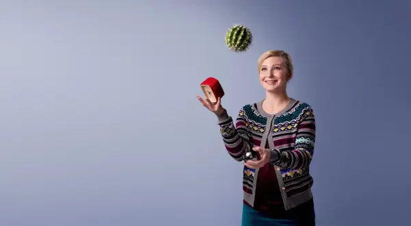 Kvinna jonglerar med en kaktus, en röd väckarklocka och en metallkula. Hon ser glad ut, som om hon klarar allt. Osa 2015.