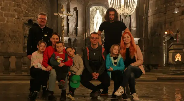 Rodzina zwiedza kopalnię soli w Wieliczce. Dzieci z rodzicami siedzą na ławce w podziemnej kaplicy, podziwiając solne rzeźby i żyrandol.