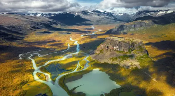 Weites Tal mit Fluss und Berg in Schwedens Nationalparks. Herbstliche Farben und dramatische Wolken über der Landschaft.