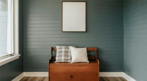 Un banc coffre en bois avec coussins, devant un mur de lambris bleu-gris fraîchement peint. Un cadre vide est accroché au-dessus.