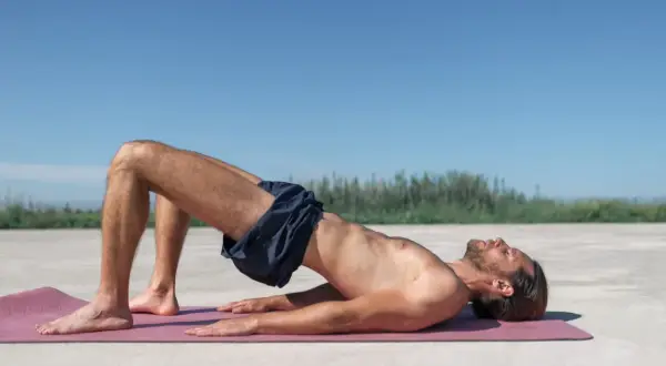 Homme pratiquant l'exercice de Kegel sur un tapis rose, le corps surélevé, les genoux pliés, sous un ciel bleu clair.