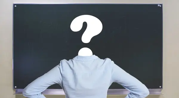 Nauczyciel z pytajnikiem zamiast głowy przed tablicą. Czy to symbol problemów z godzinami ponadwymiarowymi nauczycieli?
