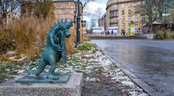 Mały smok z brązu, symbol smoczy szlak kraków, stoi na kamiennym postumencie. W tle miejski krajobraz z budynkami i drzewami.