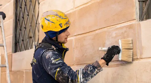 Operaio con casco giallo intento a intonacare muro vecchio con un pennello largo.
