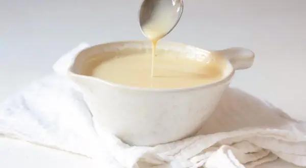 Cuchara vertiendo crema inglesa en un cuenco blanco sobre tela arrugada.
