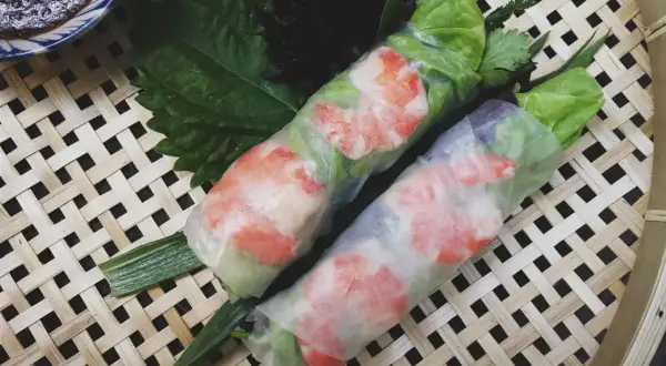 Dwa świeże sajgonki z krewetkami i warzywami, zawinięte w papier ryżowy, leżą na bambusowej macie.