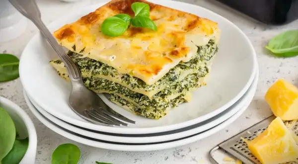 Ein Stück Spinatlasagne mit Käse überbacken, garniert mit Basilikum, auf einem Teller mit einer Gabel.