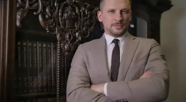 Mężczyzna w garniturze stoi przed regałem z książkami, ręce skrzyżowane na piersi.