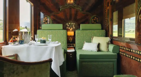 Luxuriöser Speisewagen im Orient Express: Ein gedeckter Tisch mit Obst und Champagner, grüne Sitzmöbel und kunstvolle Holzverzierungen.
